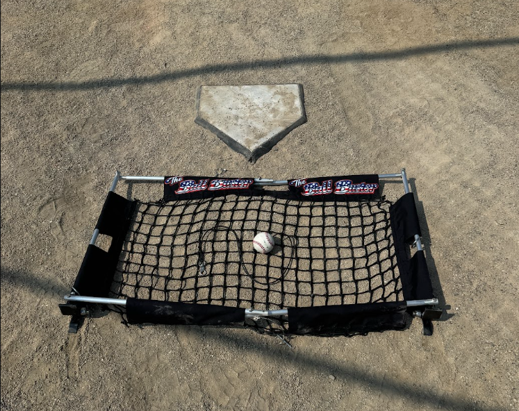 The Ball Buster hitting trainer set up at home plate — steel-and-aluminum frame with netting and a tethered ball, marked with the red-white-and-blue Ball Buster logo